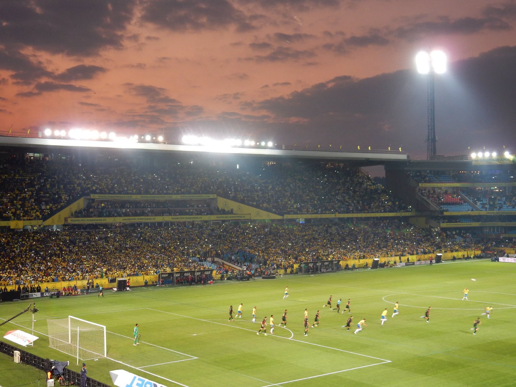 Blick auf das ausverkaufte Loftus-Versfeld-Stadion in Pretoria beim CAF-Champions-League-Finale zwischen Mamelodi Sundowns und AS FAR Rabat. Gelb gekleidete Fans füllen die Tribünen unter einem orangefarbenen Abendhimmel.