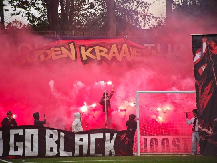 HBS-Fans zünden im Derby gegen Quick roten Rauch und mehrere Bengalos hinter dem Tor. Über dem Block hängt ein großes Banner mit der Aufschrift „Den Kraaien“.