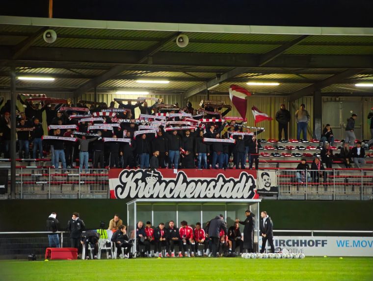 Fans der Sportfreunde Siegen halten rot-weiße Schals im Gästeblock des Hemberg-Stadions in Iserlohn hoch. Vor der Tribüne hängt die Zaunfahne „Krönchenstadt“.