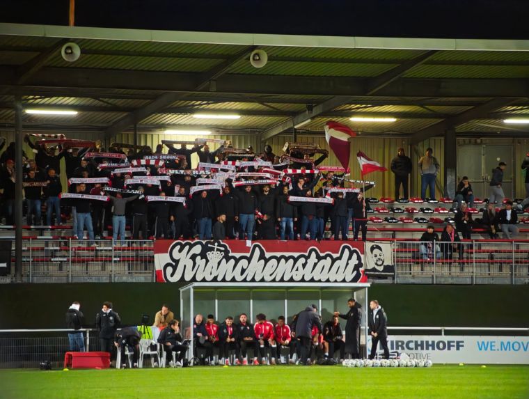 Fans der Sportfreunde Siegen halten rot-weiße Schals im Gästeblock des Hemberg-Stadions in Iserlohn hoch. Vor der Tribüne hängt die Zaunfahne „Krönchenstadt“.