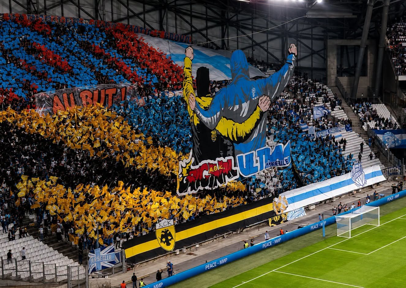 Freundschaftschoreografie der Ultras von Olympique Marseille und AEK Athen beim Europa-League-Duell im Stade Vélodrome. Auf der Tribüne sind eine große Blockfahne mit zwei sich umarmenden Fans sowie gelbe, blaue und rote Papptafeln zu sehen.