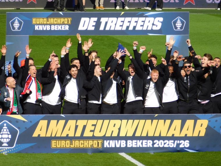 Spieler und Betreuer von De Treffers jubeln gemeinsam auf dem Spielfeld und halten die blaue Trophäe hoch. Vor ihnen steht ein großes Schild mit der Aufschrift „Amateurwinnaar KNVB Beker 2025/26“.