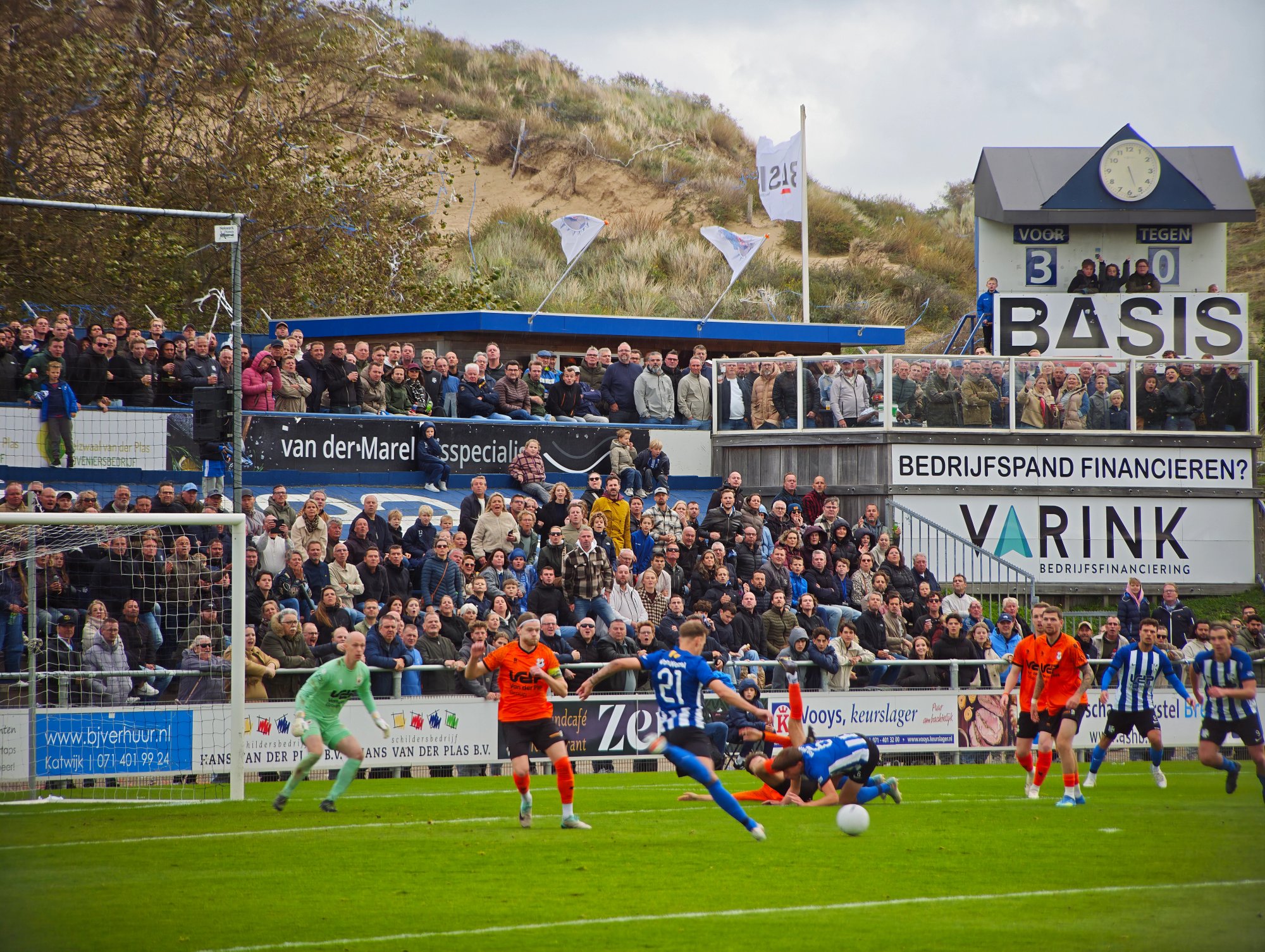 Spielszene beim Derby Quick Boys gegen VV Katwijk im Sportpark Nieuw Zuid. Im Hintergrund stehen zahlreiche Zuschauer auf den dicht gefüllten Tribünen vor der typischen Dünenlandschaft von Katwijk.