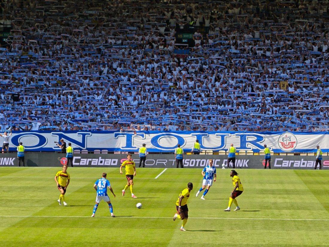 Großer Hansa-Rostock-Gästeblock mit Banner und hochgehaltenen Schals hinter dem Spielfeld in Watford.