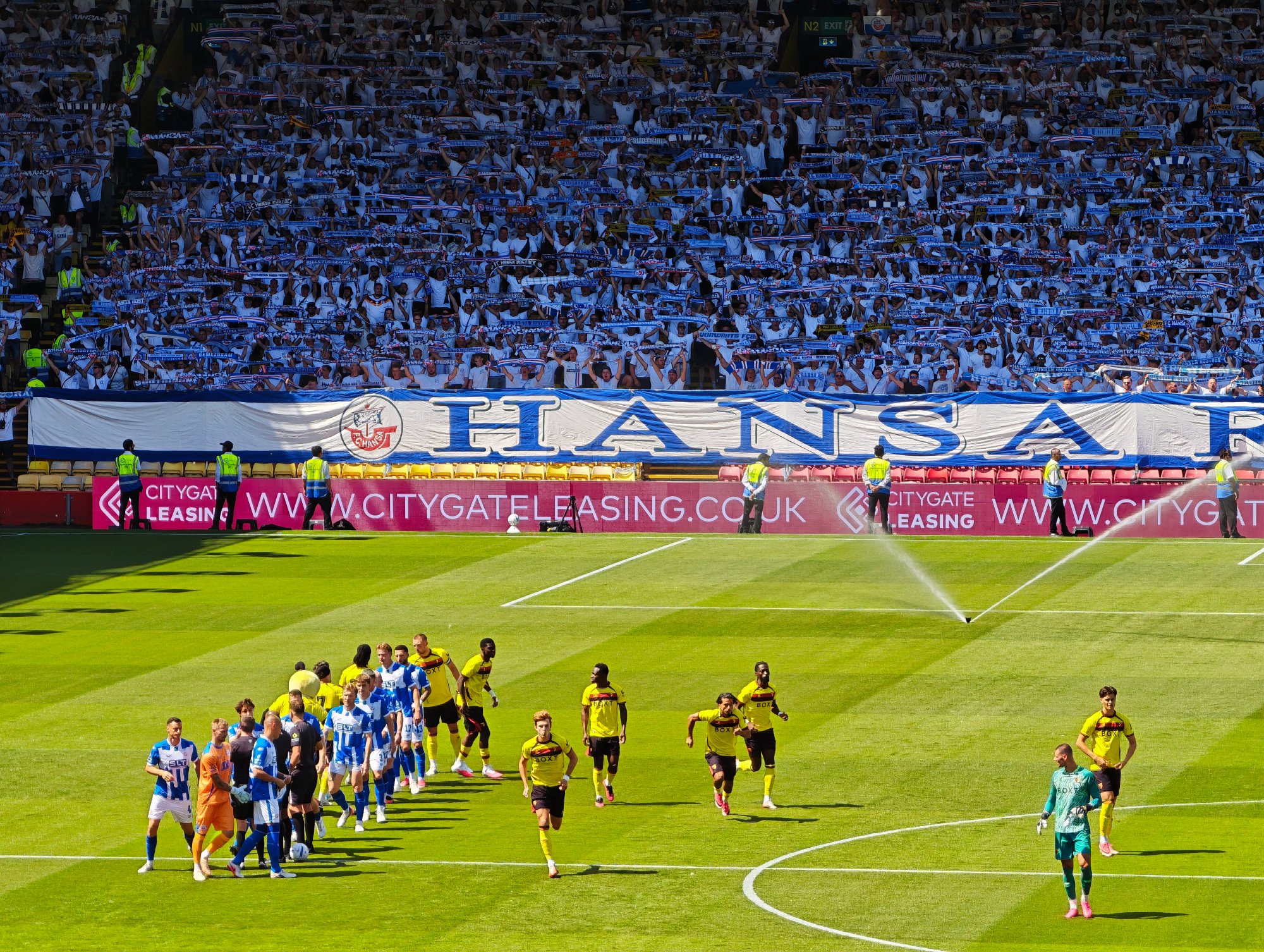 Hansa-Rostock-Fans mit hochgehaltenen Schals und großem „On Tour“-Banner hinter dem Tor in der Vicarage Road.