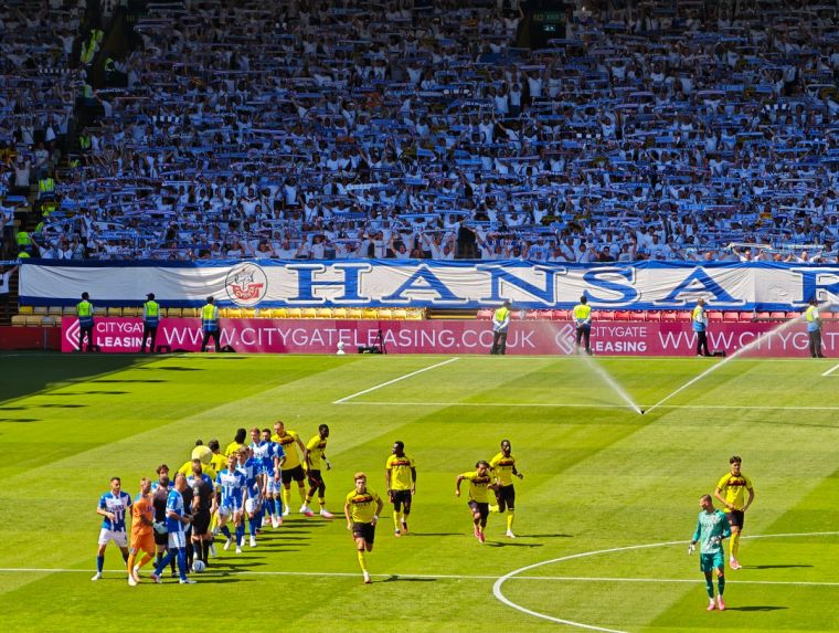 Hansa-Rostock-Fans mit hochgehaltenen Schals und großem „On Tour“-Banner hinter dem Tor in der Vicarage Road.