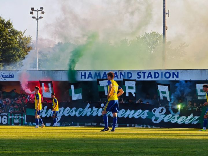 Spielszene vor der überdachten Tribüne im Hamalandstadion, dahinter farbiger Rauch und Fanaktion von Preußen Münster.