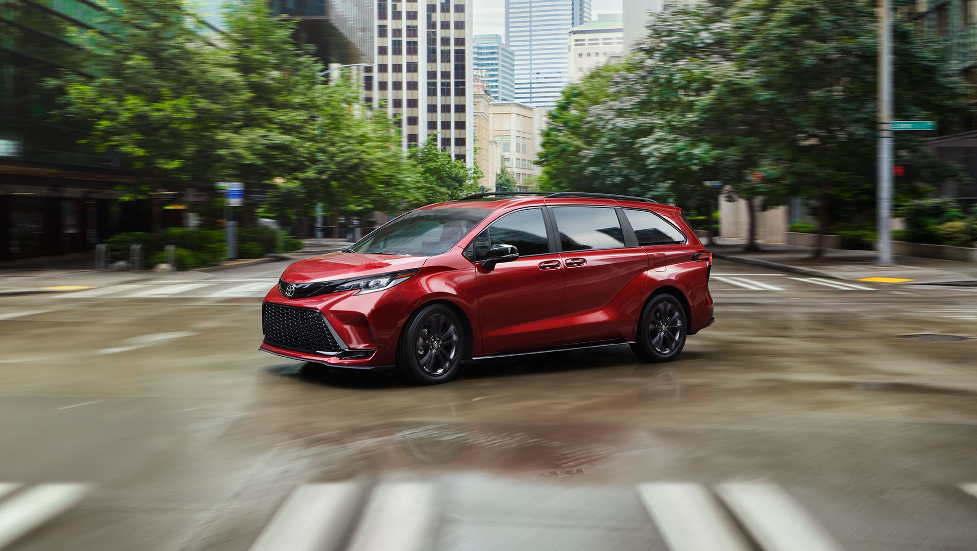 A red minivan drives through an empty, wet city intersection surrounded by tall buildings and greenery on a cloudy day.