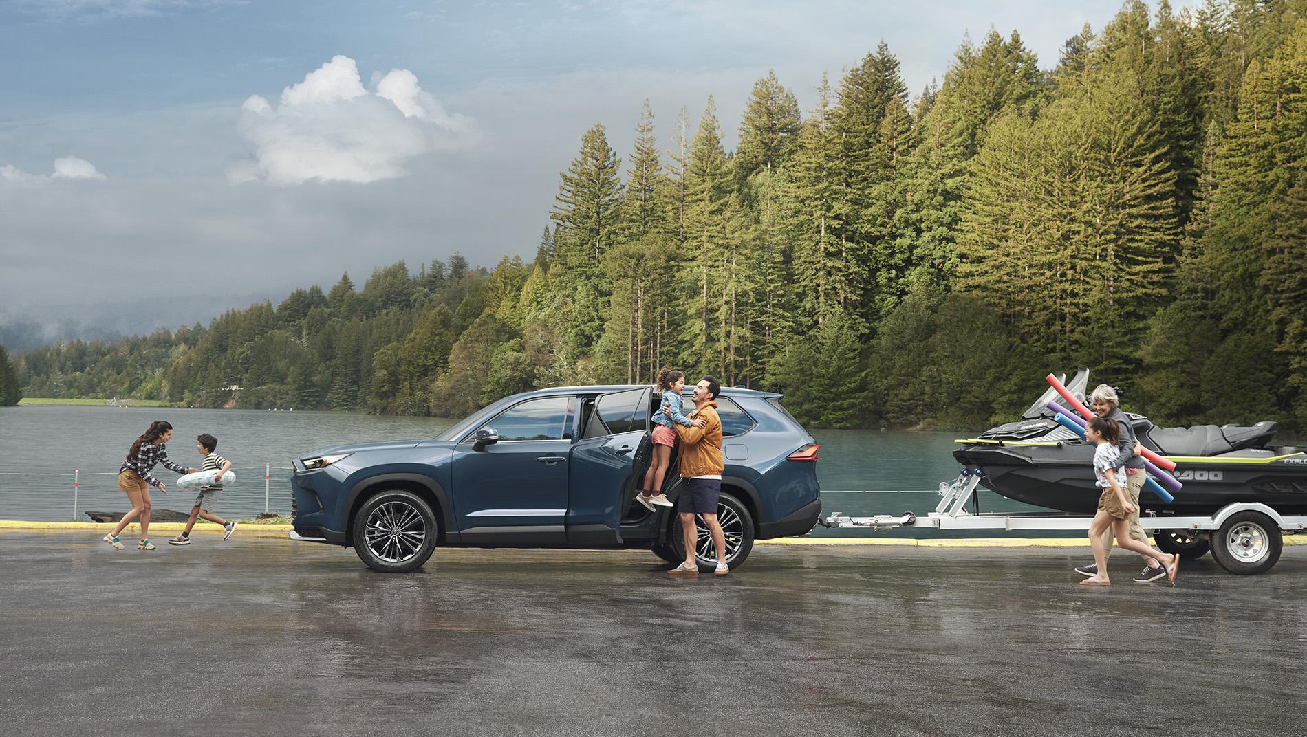 Toyota SUV towing 2 jet skis in front of a lake that is surrounded by evergreen trees. Family getting out of the vehicle while kids play.