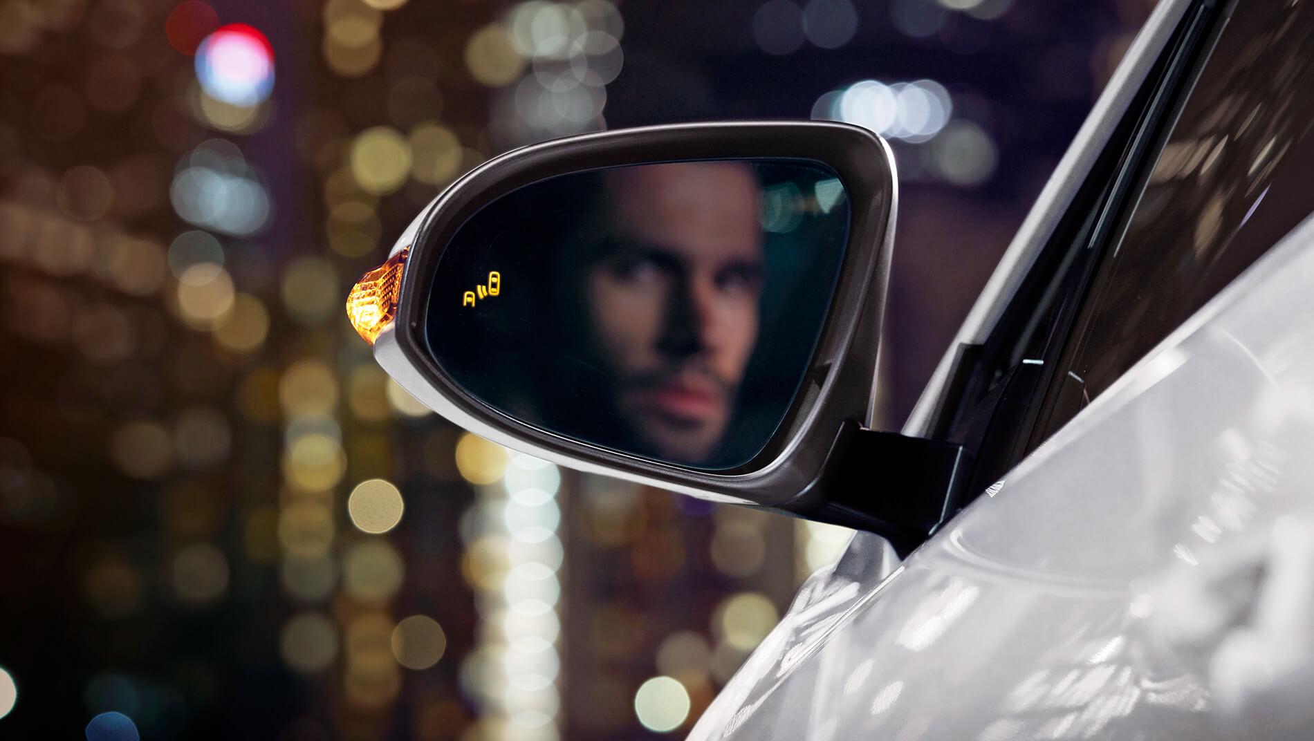 mans face in side mirror of a white Toyota C-HR at night with city lights beyond