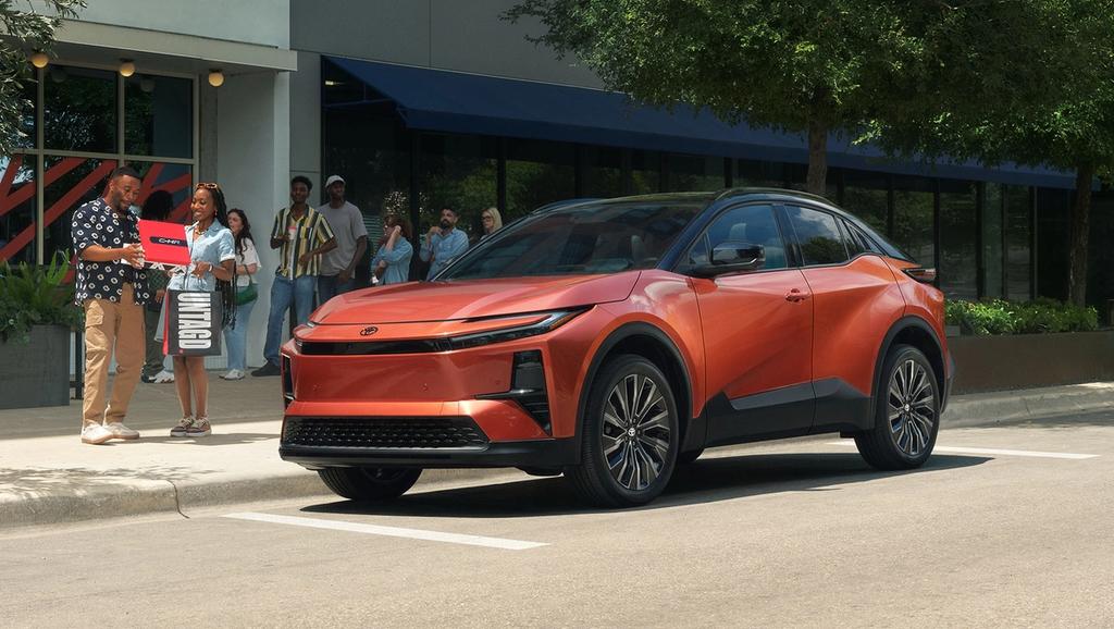 A sleek, orange electric SUV is parked on a city street near a sidewalk. Two people stand beside it, one holding a red box, while others are gathered in the background near a building with large windows.