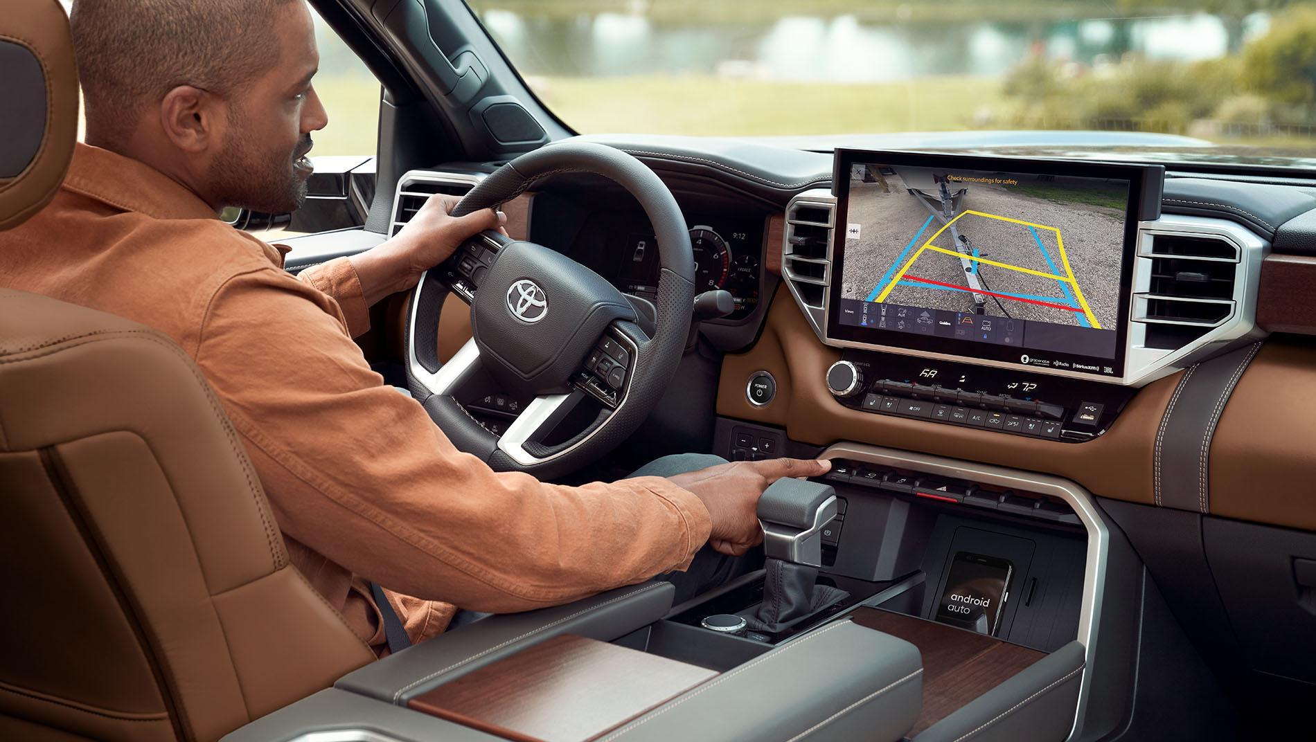A person seated in a 2024 Toyota a Tundra is using the gear shift while looking at a large touchscreen display on the dashboard.