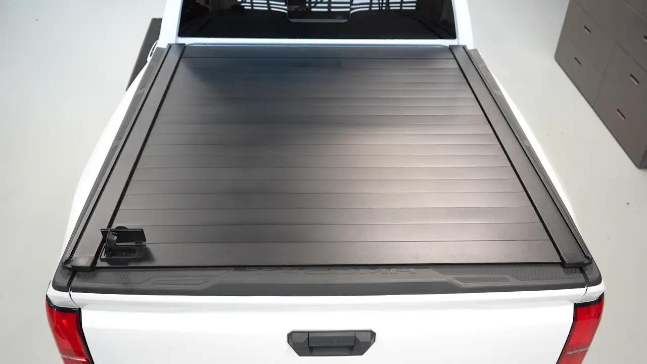 A white pickup truck with a closed, black, hard retractable tonneau cover over the bed, viewed from above and behind in a garage setting.