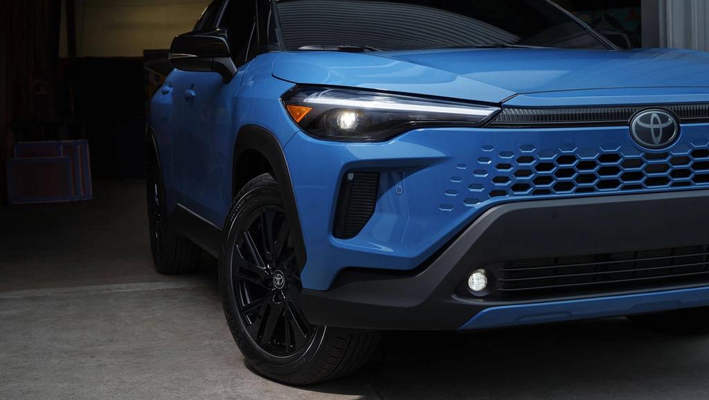 Close-up of the front side of a blue Toyota SUV parked indoors, showing the headlight, front grille with honeycomb design, Toyota badge, and black alloy wheels.