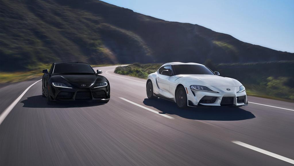 2 Toyota GR Supras, 1 black and 1 white, driving down a curve along a mountain road