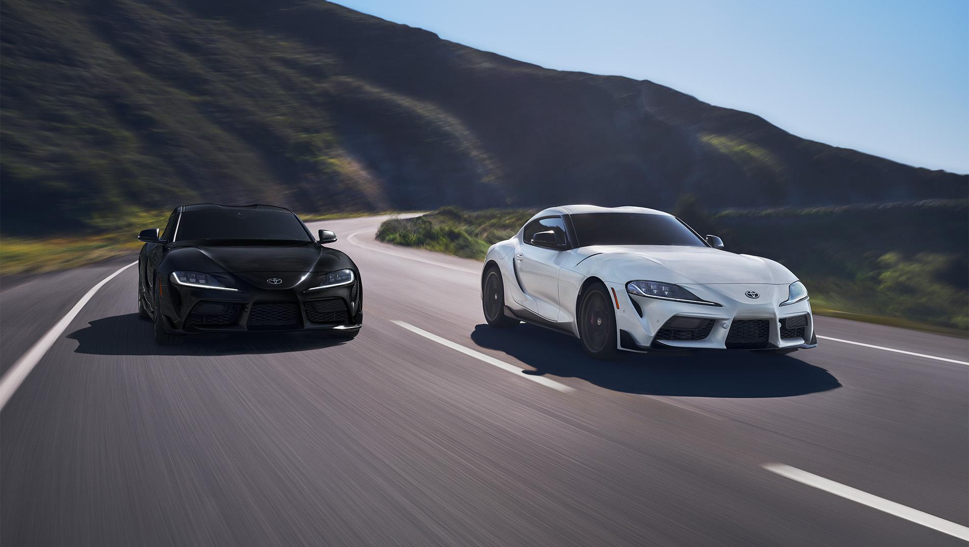 2 Toyota GR Supras, 1 black and 1 white, driving down a curve along a mountain road