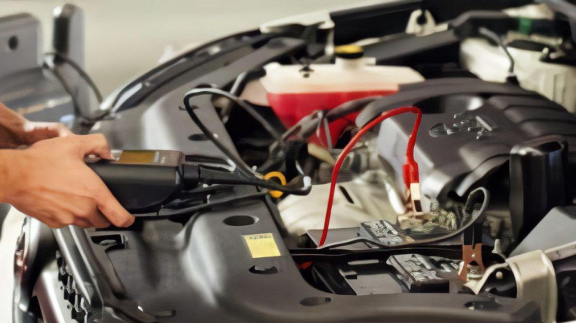 A person uses a battery tester with connected cables to check a car battery under an open hood, with engine components and wires visible.A person uses a battery tester with connected cables to check a car battery under an open hood, with engine components and wires visible.