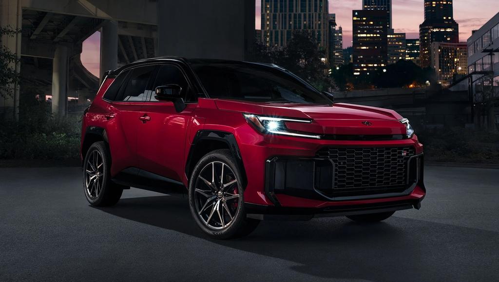 A shiny red SUV with black accents is parked on a city street at dusk, with tall buildings and lights in the background. The car features bold styling and large alloy wheels.
