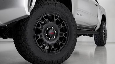 Close-up view of a rugged black off-road tire and wheel on a silver pickup truck, showing the tire tread and part of the vehicle's body. The background is a clean, light-colored floor and wall.