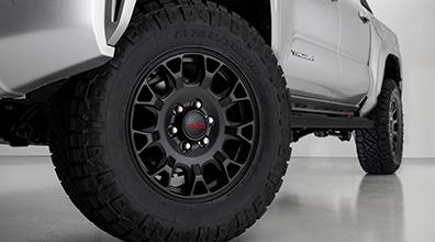 Close-up view of a rugged black off-road tire and wheel on a silver pickup truck, showing the tire tread and part of the vehicle's body. The background is a clean, light-colored floor and wall.