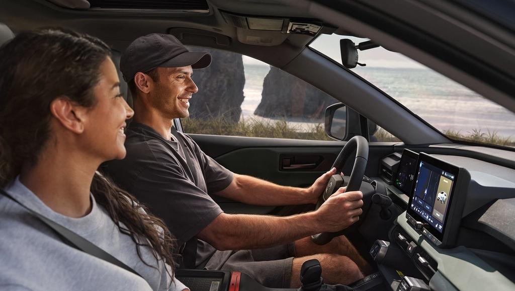 A man and a woman smile while sitting in the front seats of a car parked near a scenic beach, with cliffs and the ocean visible through the windows. The car's dashboard and touchscreen display are in view.