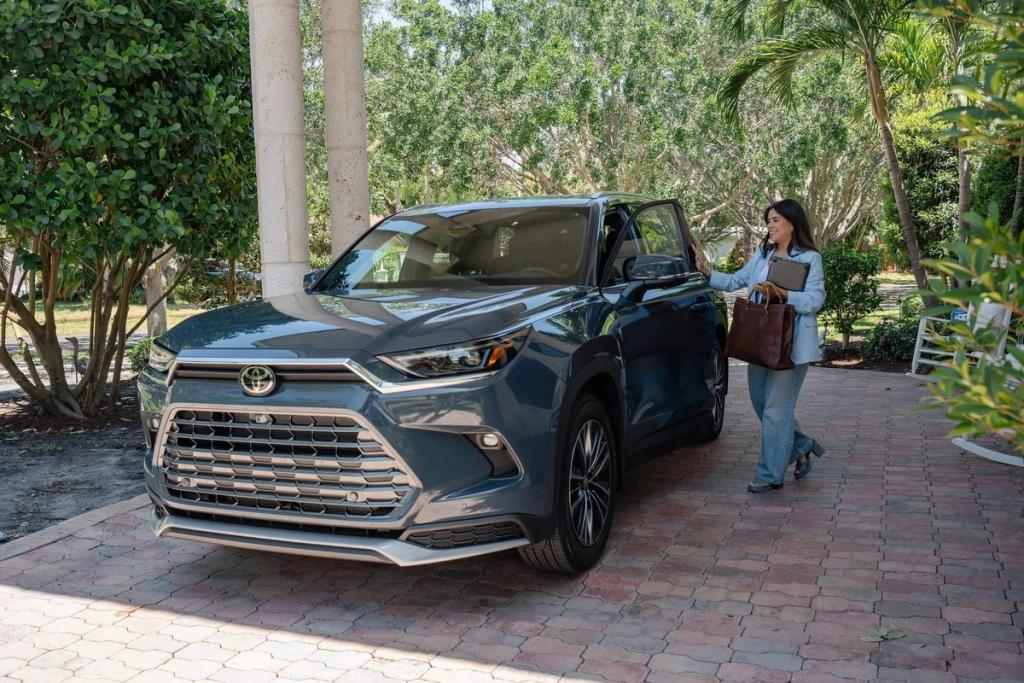 A woman holding a brown bag and laptop approaches the driver's side of a parked blue Toyota SUV on a sunlit driveway, surrounded by green trees and plants.