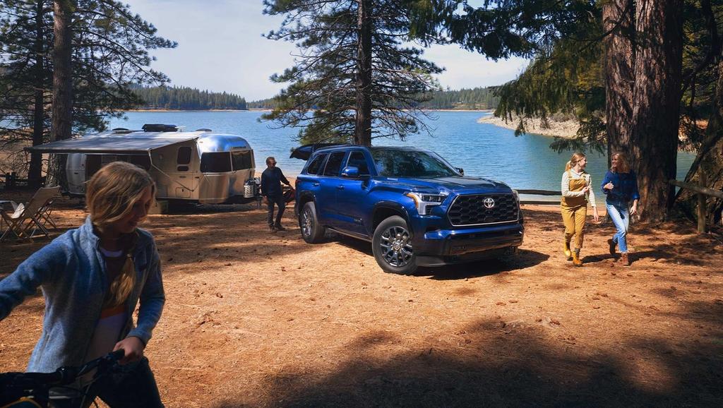 A blue 2024 Toyota Sequoia i-FORCE MAX Hybrid is parked near a shiny trailer at a lakeside campsite surrounded by trees. Four people are engaging in outdoor activities.