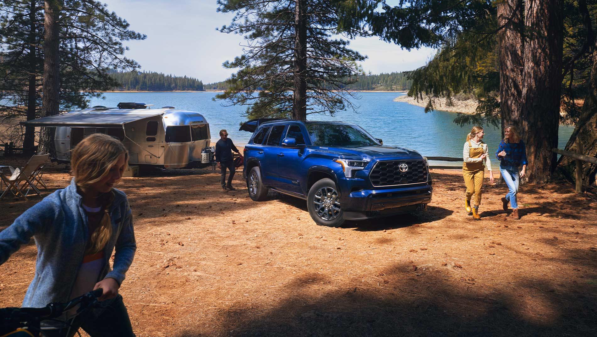 A blue 2024 Toyota Sequoia i-FORCE MAX Hybrid is parked near a shiny trailer at a lakeside campsite surrounded by trees. Four people are engaging in outdoor activities.