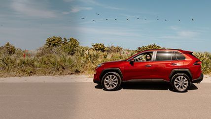 A red toyota highlander is parked on a road beside a sandy beach while a woman is inside the vehicle.