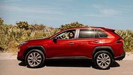 A red toyota highlander is parked on a road beside a sandy beach while a woman is inside the vehicle.