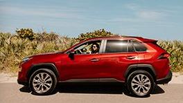 A red toyota highlander is parked on a road beside a sandy beach while a woman is inside the vehicle.