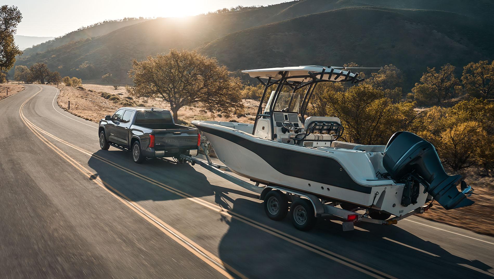 A 2024 Toyota Tundra towing a large boat on a trailer down a scenic, winding road surrounded by hills.