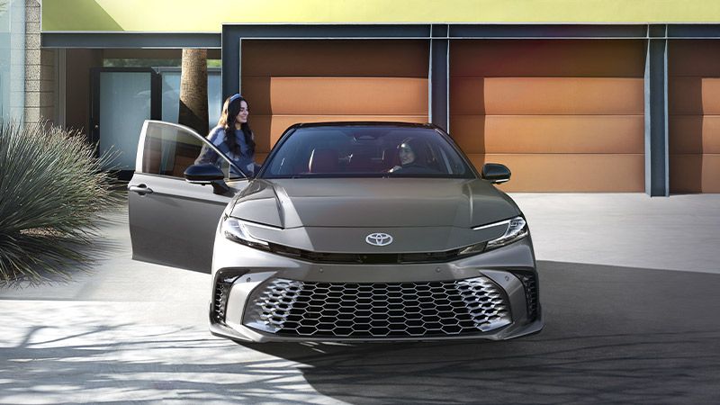 A woman stands next to an open driver’s door of a gray Toyota sedan parked in front of a modern house with three brown garage doors.