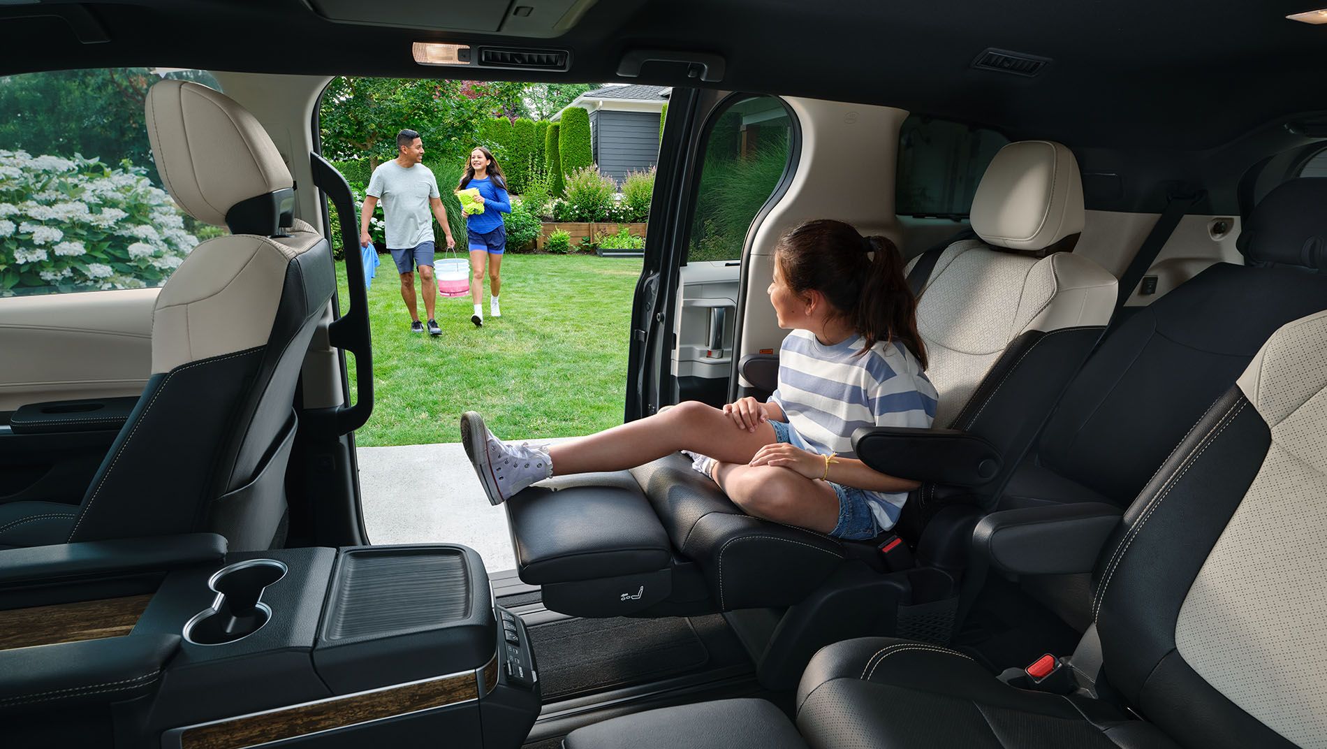 A girl sits inside a minivan with the sliding door open, looking at a family of three—two adults and one child—walking towards the vehicle in a suburban neighborhood with green lawns and flowers.