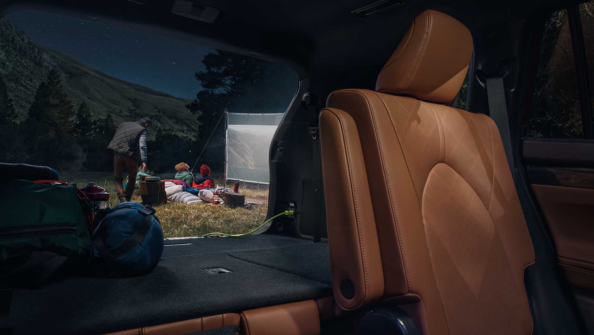 View from inside a car with tan leather seats, looking out at a nighttime campsite where three people sit near a tent and a campfire