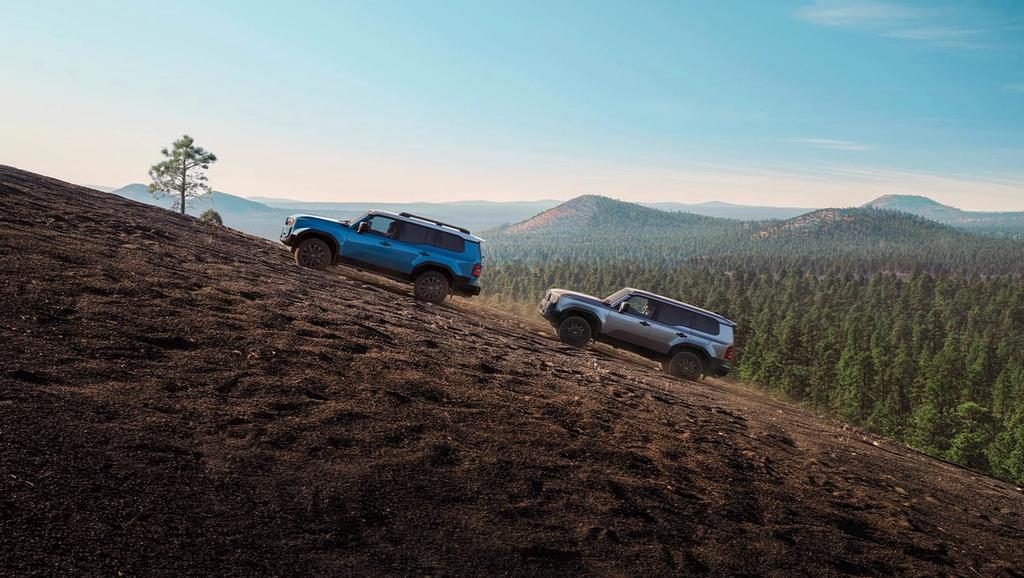 blue 2025 Toyota Land Cruiser hybrid climbing a mountain hill followed by an older generation Land Cruiser