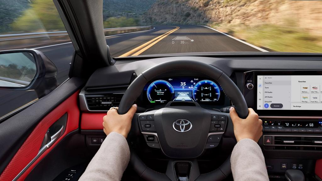 A person drives a Toyota car on a curving road; the dashboard displays digital speed and navigation, while a screen on the right shows music and map options.