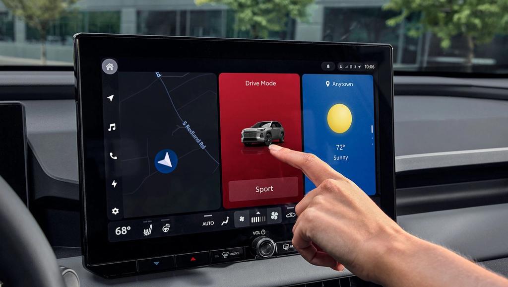 A hand touches a car’s touchscreen display showing navigation, drive mode set to Sport with a car image, and weather info for Anytown showing sunny and 72°F. Trees and buildings are visible through the windshield.