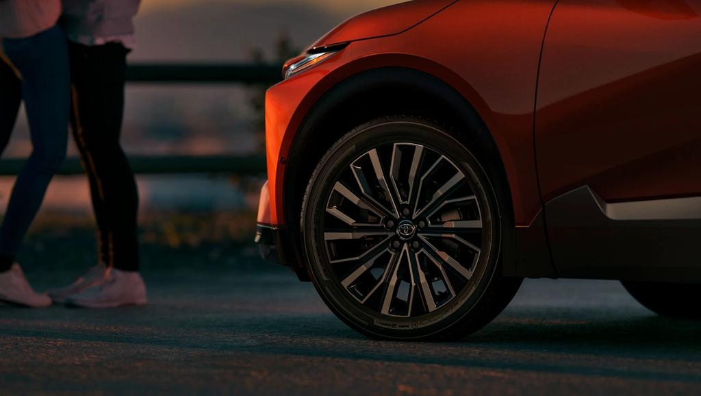 Close-up of the front wheel and fender of an orange car parked on a road at sunset, with two people standing in the blurred background.