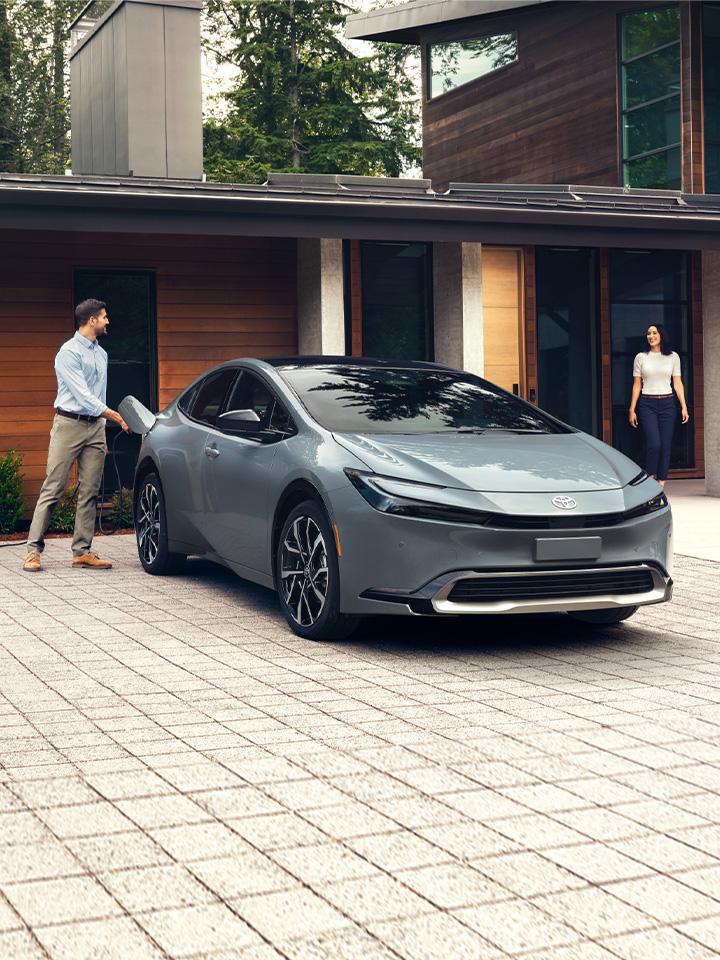 grey 2024 Toyota Prius Prime Plug In Hybrid parked in front of a house with a man unplugging it before he and a woman get in