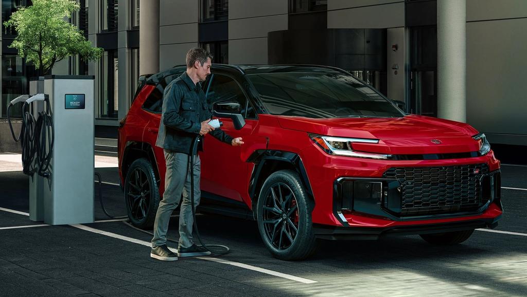 A person stands next to a red electric SUV, holding a charging cable and plugging it into the vehicle at a charging station in an urban setting.