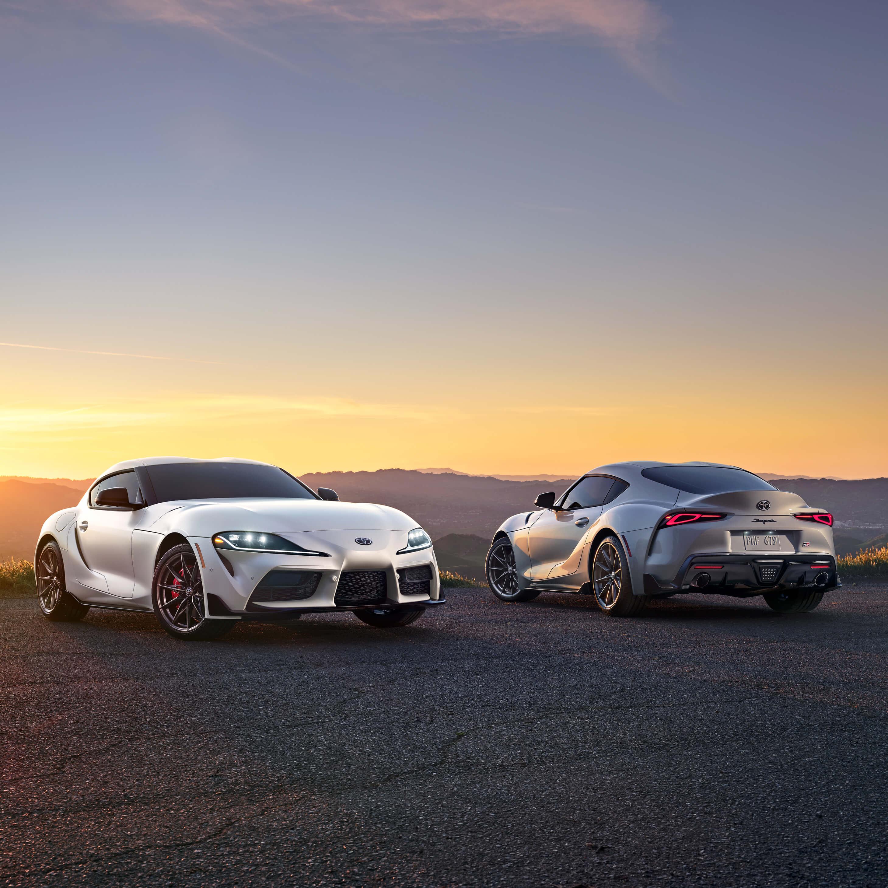 2 Toyota Supra sports cars parks on a hill overlooking a sunset on the mountains