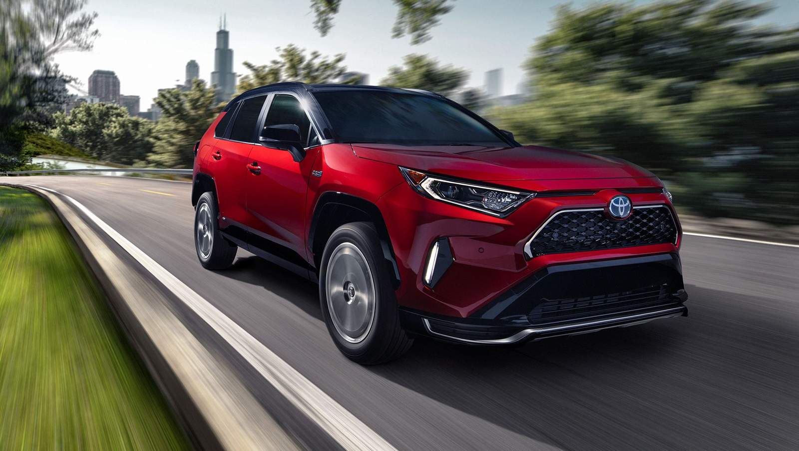 red Toyota RAV4 Prime driving down tree lined road with a city in the background