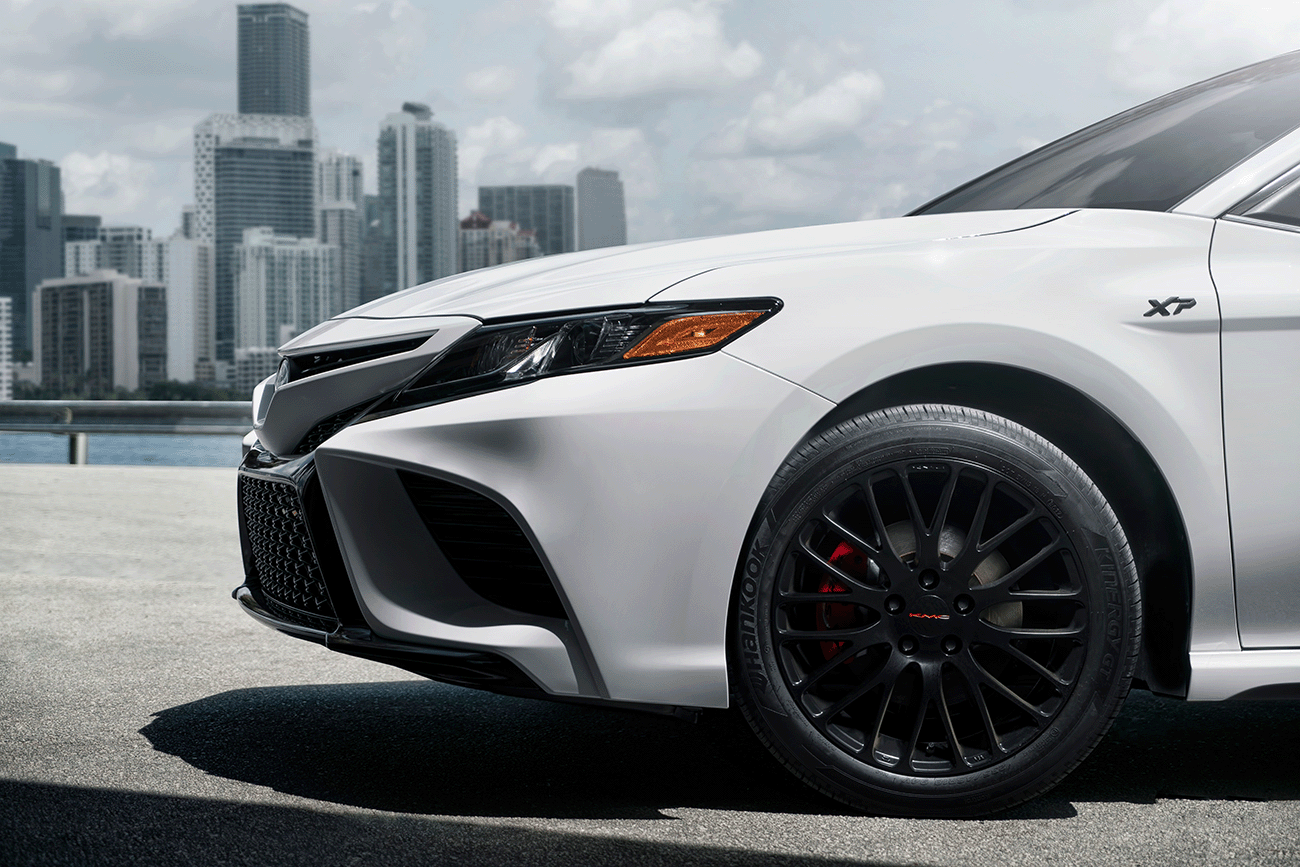 close up image of the front of a white Toyota Camry XP XSeries upgraded accessories package showing the upgraded XSeries wheels and black gloss emblem