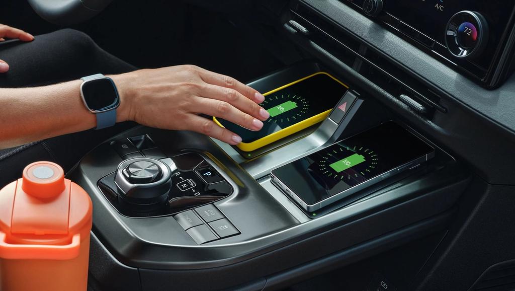 A person’s hand places a smartphone on a wireless charging pad in a car. Another phone is also charging. The car console has controls and an orange water bottle is nearby. Both phones show charging icons on their screens.