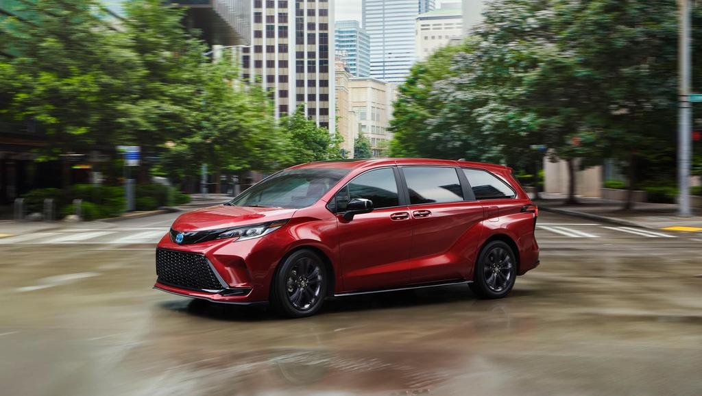 A red toyota 2025 sienna driving on a wet city street with blurred buildings and trees in the background.