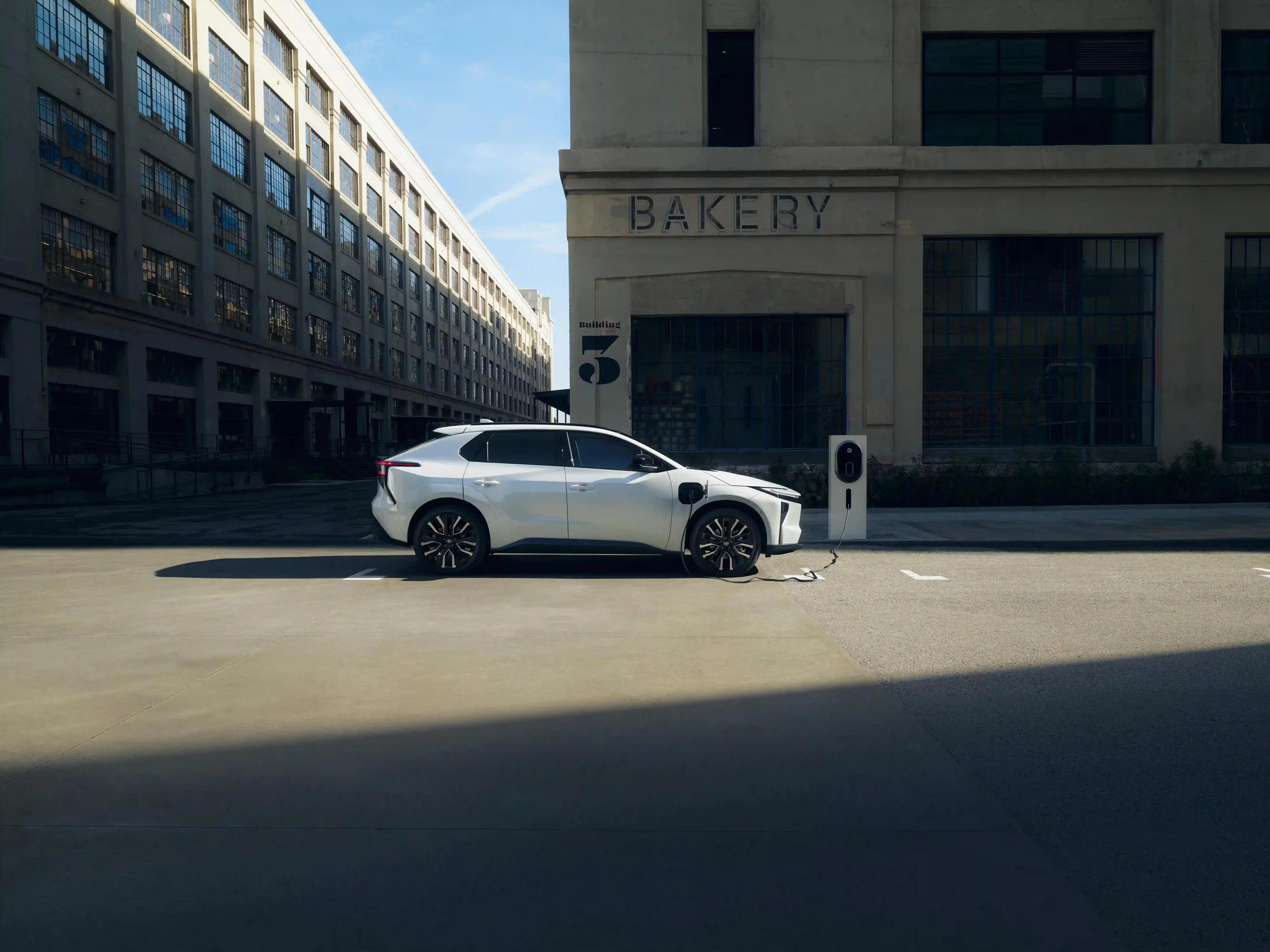 A white electric SUV is parked and charging at a charging station in front of a large industrial-style building with "BAKERY" written above the entrance. The scene is sunlit, with deep shadows on the pavement.