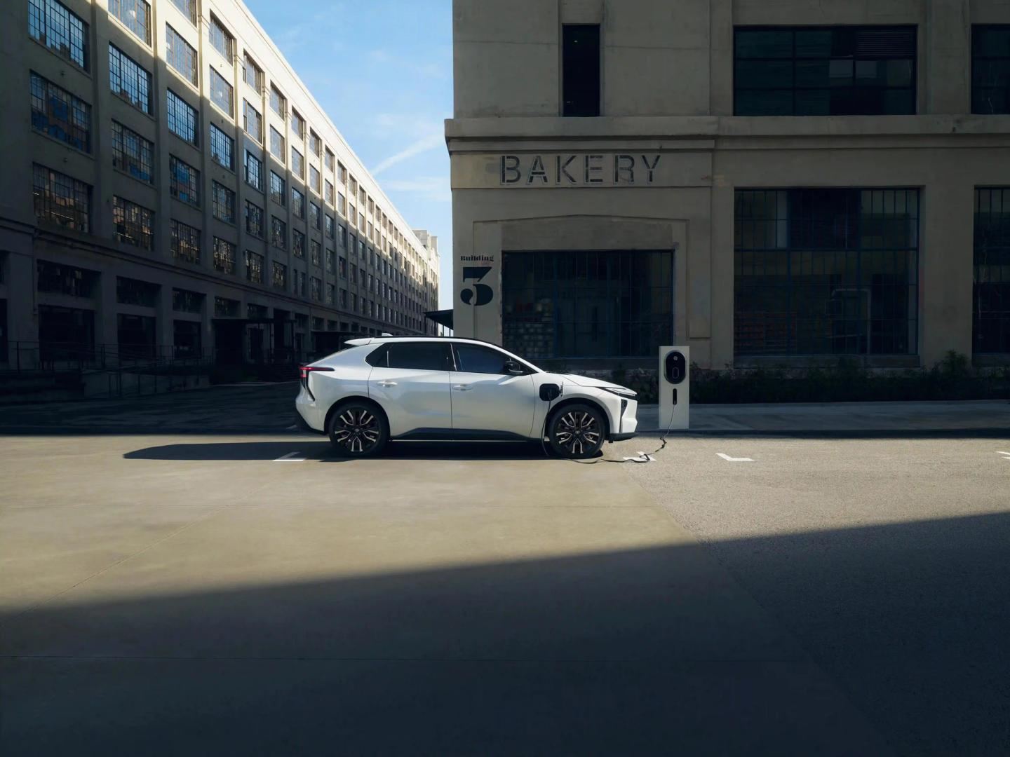 A white electric SUV is parked and charging at a charging station in front of a large industrial-style building with "BAKERY" written above the entrance. The scene is sunlit, with deep shadows on the pavement.