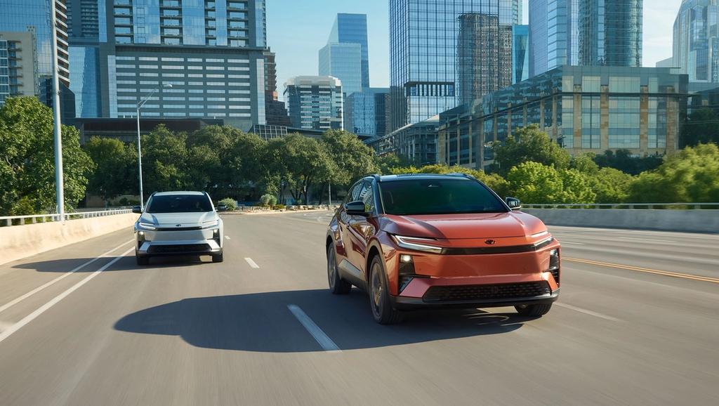 Two modern electric SUVs drive on a city street with tall glass buildings and trees in the background on a sunny day. The SUV in front is orange, while the one behind is white.