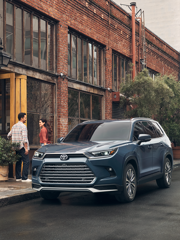 Blue 2024 Toyota Grand Highlander Hybrid parked on a street. a man and a woman are walking into a storefront